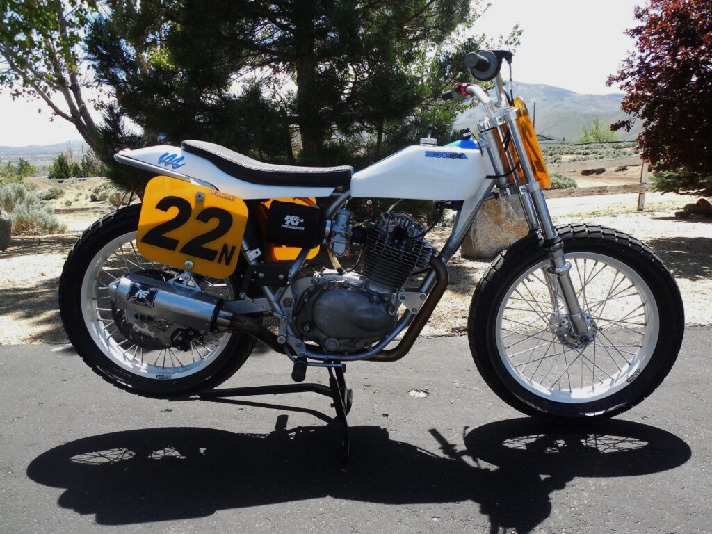 Flat track motorcycle with a yellow number 22 plate, white tank and black seat, standing in the sun